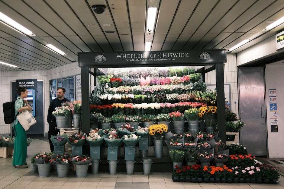 A vibrant floral display at Wheeler of Chiswick, situated in an underground station, showcasing an assortment of fresh flowers arranged in metal buckets and wrapped bouquets. The display includes sunf