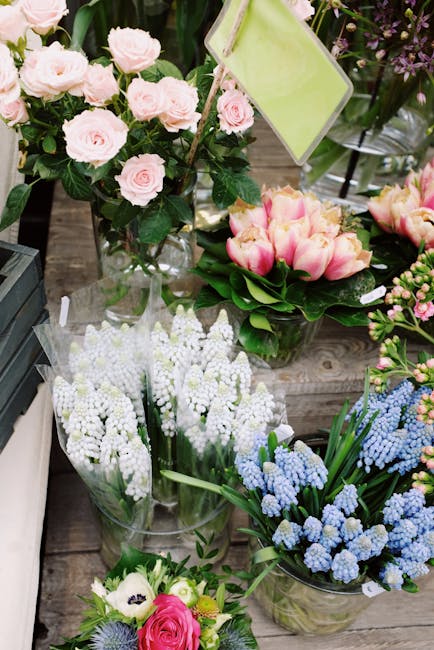A variety of fresh floral arrangements displayed on a wooden surface, including a bouquet of pale pink roses with dark green foliage in a glass vase, a cluster of pink and cream tulips with green leav